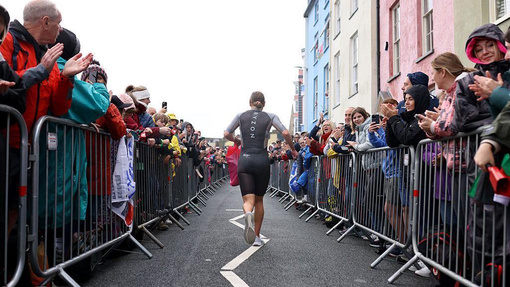 A runner runs away from the camera wearing black top and short with spectators standing either side of the road behind barriers