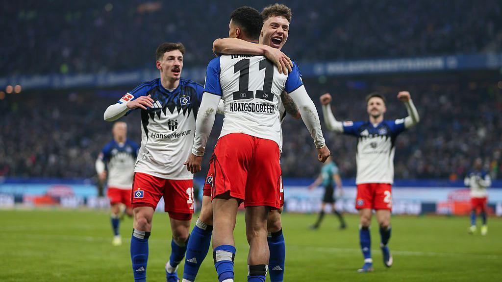 Hamburger SV players celebrate a goal against Union Berlin. They are wearing a blue and white kit with red shorts and a black trim.
