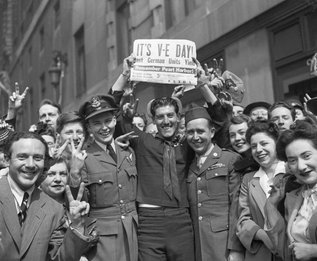 A photo from 1945 showing a happy group of civilians and soldiers holding a newspaper with the headline "It's VE Day!"