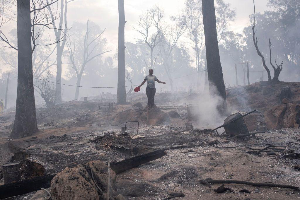 A woman stands among the burnt remains, surrounded by smoke and bare trees.