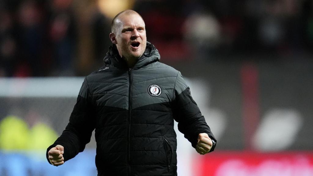 Liam Manning wearing a black and grey puffer jacket with a Bristol City badge on the chest. He is pumping his fists at his sides.