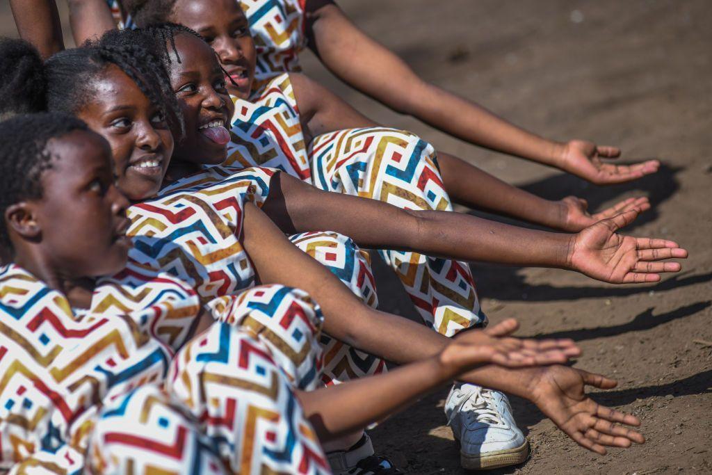 Girls in matching multicoloured dressed sit in a row on the ground with with arms outstretched and smiling - Nairobi, Kenya, Saturday 7 December 2024