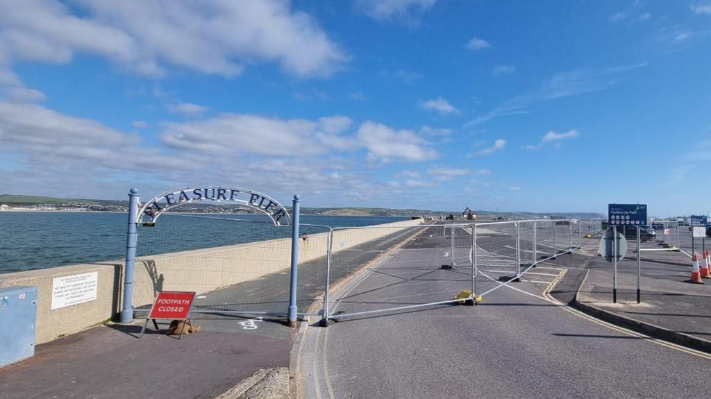 The road on to Weymouth Peninsula car park blocked off with Heras fencing. There is a red sign on the pavement saying footpath closed. To the left of the picture is the sea and Weymouth Bay.