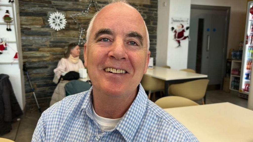 A man sits at a table in a coffee shop. He is smiling, and is bald. He has short grey hair and is wearing a checked blue shirt. Behind him are a number of tables and chairs. A woman is sitting at a table in the background