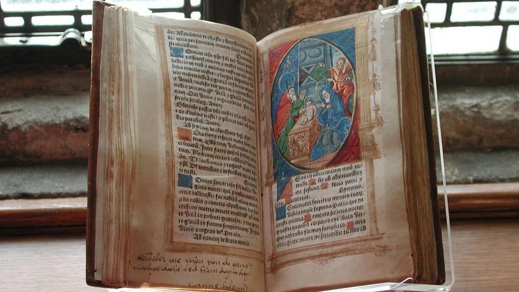 The Book of Hours prayer book signed by Anne Boleyn. It is an old book, open on a page with text and an image on the right page. It is stood in a window.