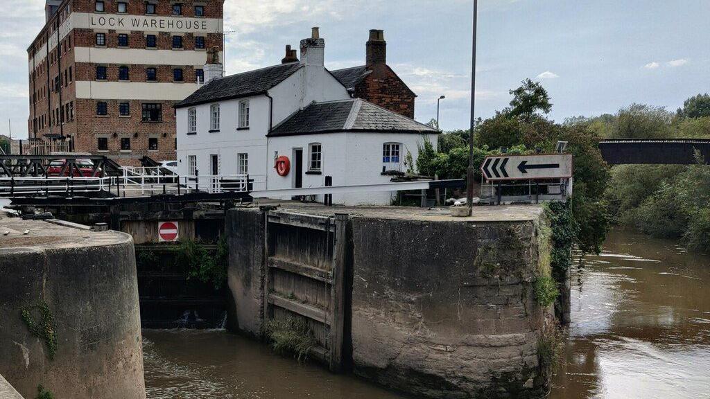Gloucester Lock. To the right is a white lock house building and behind that a brick building marked "lock warehouse". The canal can be seen in the foreground and to the right. The water level appears to be very low.