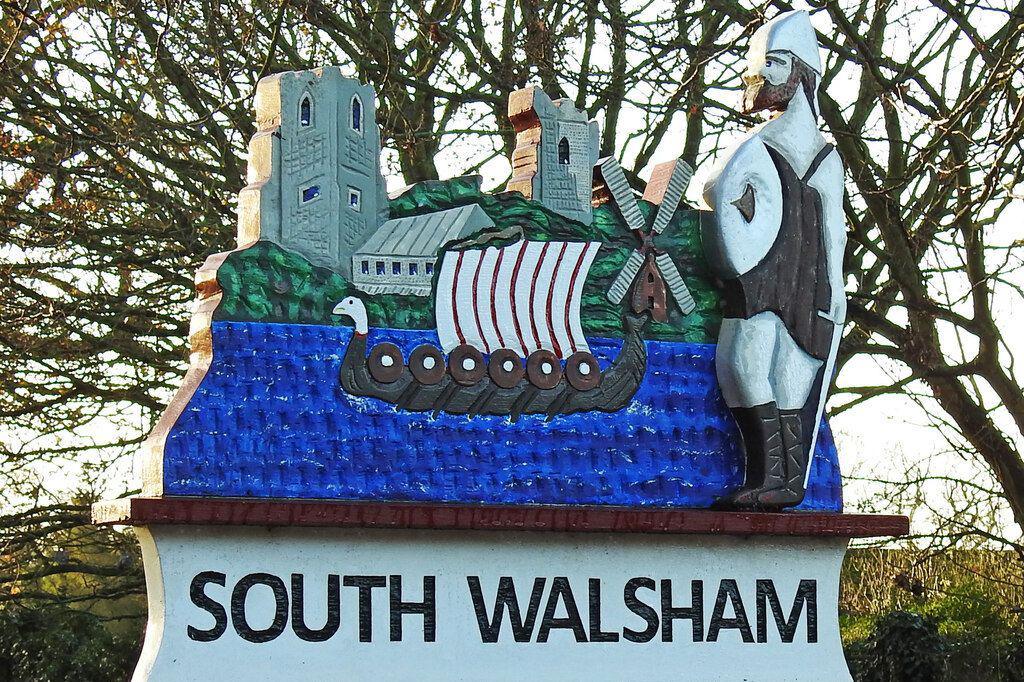 We can see the village sign of South Walsham. In the centre is a Viking longship sailing on water and to the right is an armoured Viking warrior. In the background we can see a church tower, windmill and other buildings.