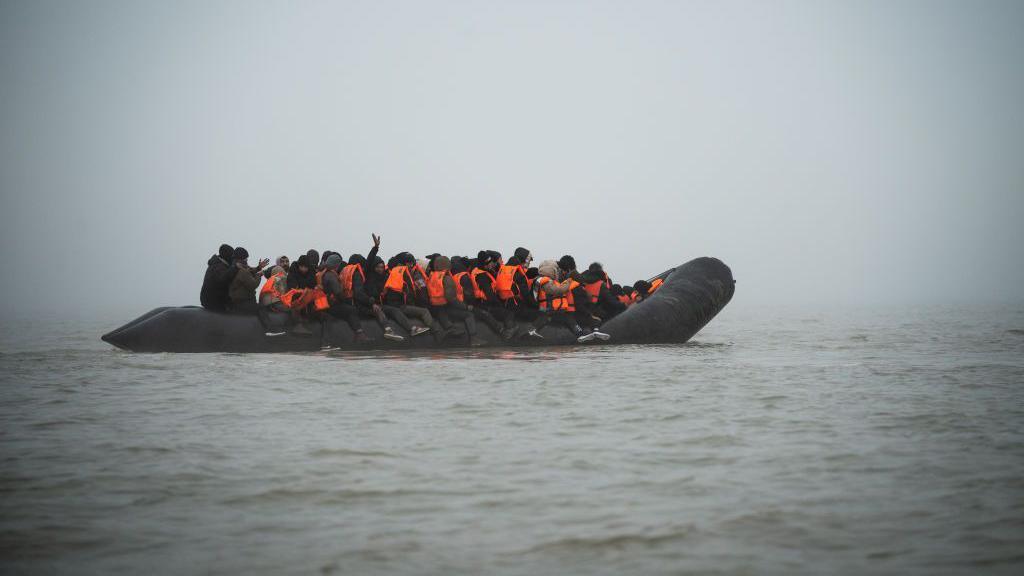 A black rubber dinghy filled with passengers, most wearing orange life vests, floats in fog on the English Channel. One person raises their hand.