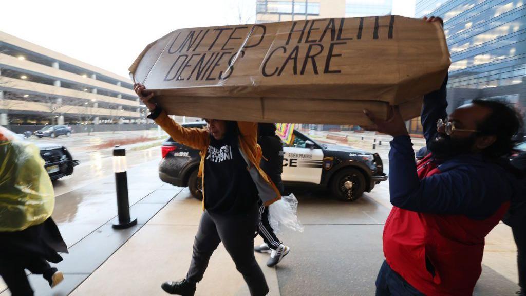 Two people in the midst of a protest holding a coffin which says "united health denies care" on it