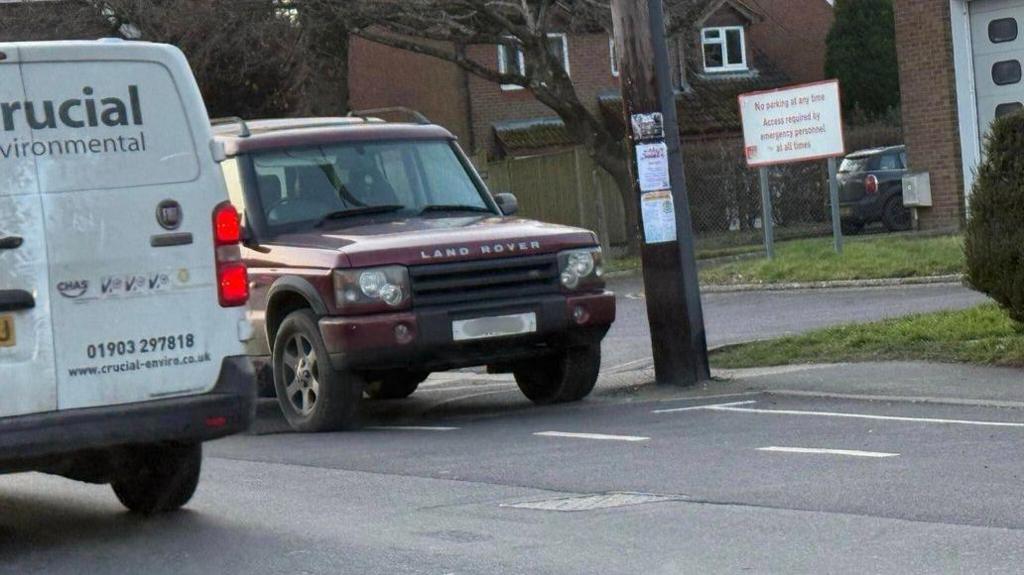 A red land rover parked on a residential road in front of a sign that says "no parking at any time. Access required by emergency personnel at all times". It is blocking an entrance. A white van driving past it is also in shot. 