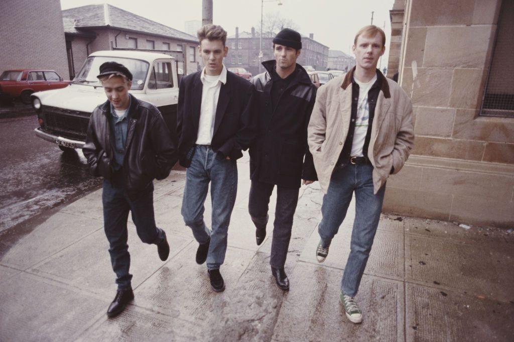 The four members of Wet Wet Wet walk along the street in Glasgow, four abreast in 1987. It's a typical Glasgow street with tenement flats in the background, cars parked on wither side of the street. The four wear casual clothes and all wear jeans. It is a drizzly day.