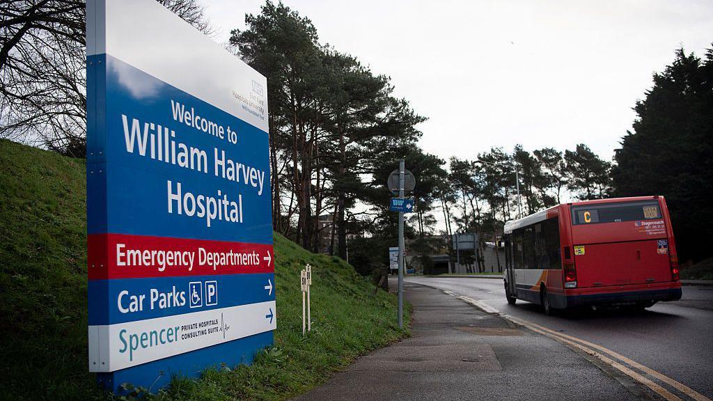 A blue sign by a roadside, on a grass hill. The sign reads Welcome to William Harvey Hospital and has directions to emergency departments and car parks underneath.