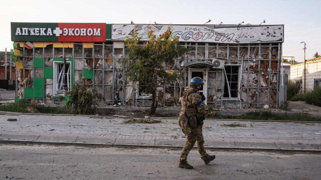 A soldier in fatigues and a helmet walks past a dilapidated building