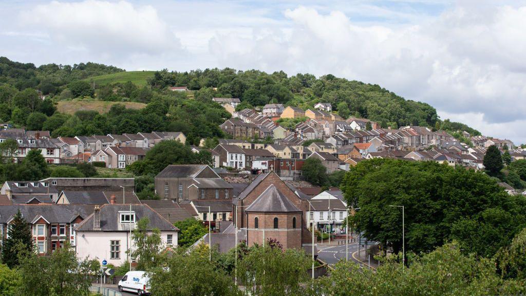 An view of Pontypridd. There are rows of terraced houses sprawled below a hill which is mostly covered in green trees