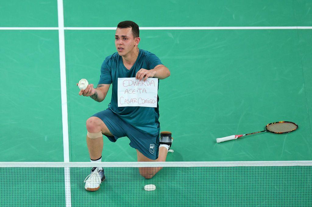 rogerio looks anxious as he bends down on one knee on the badminton court with his racket behind him and a ring in his hand looking out to his girlfriend who is not seen in this image