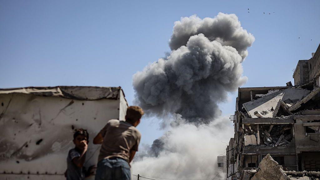Two boys, one smaller than the other, look at a huge mushroom cloud of smoke following an Israeli strike. To their right is a badly damaged building. They are sheltering behind what appears to be a a metal container.