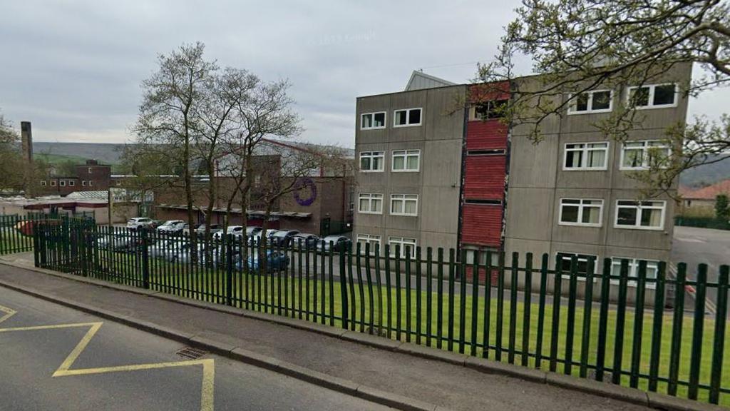 External view of The Valley Leadership Academy in Bacup which has a black fence and a handful of trees at the front of the school and car park to the right with hills in the distance.