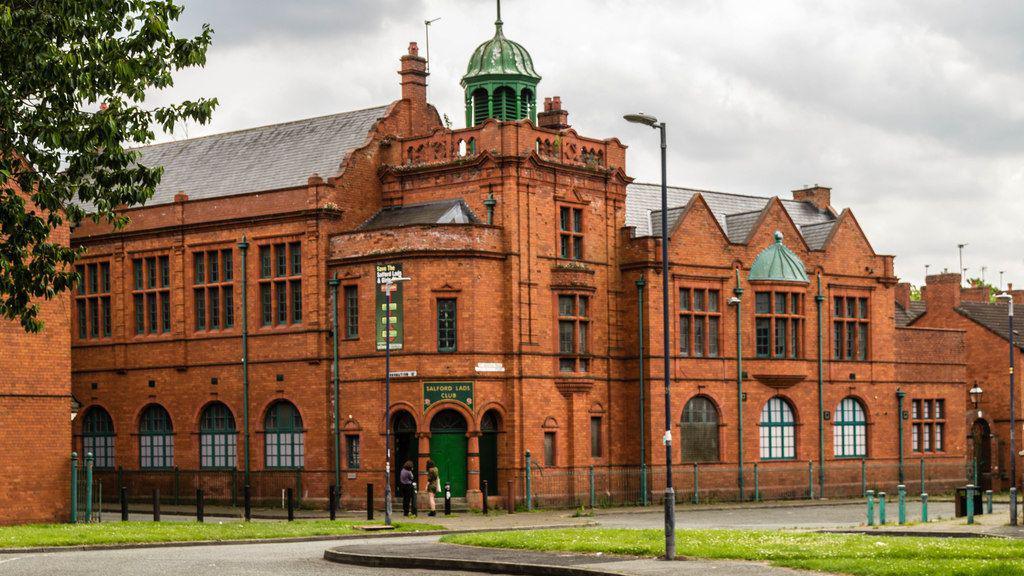 The red brick Salford Lads Club building in Orsdall, with a street lamp, road and grass verges seen in front of the club on an overcast day. 