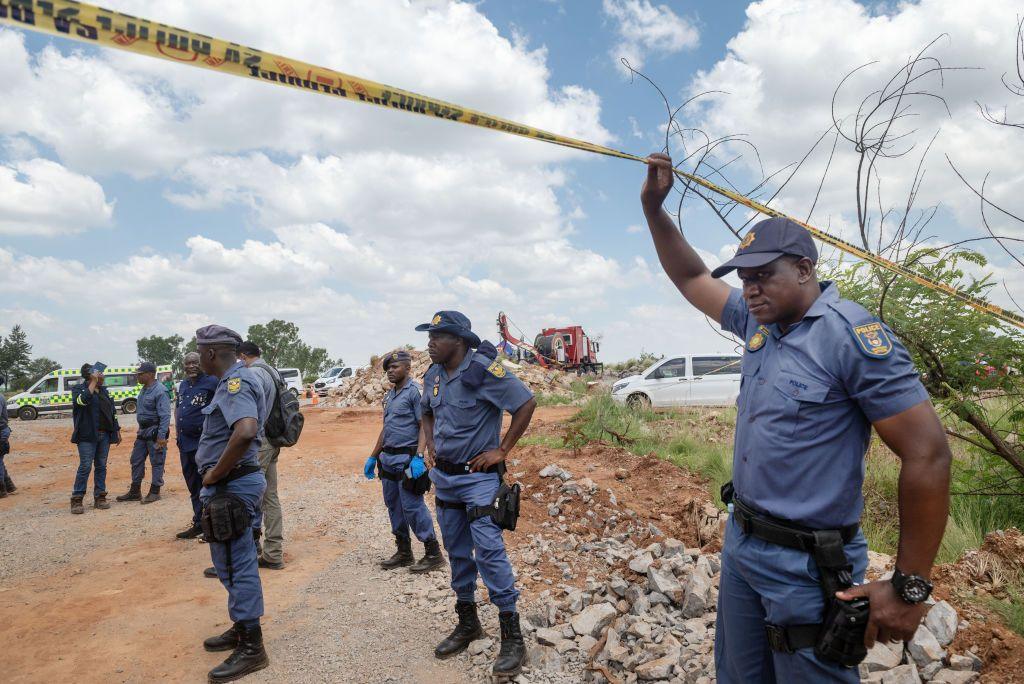 Policemen stand on a dirt road as one in the foreground lifts his arm up to hold some police tape aloft.