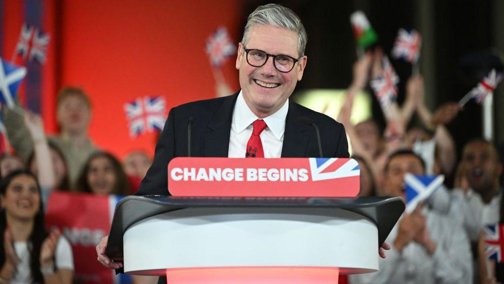 Sir Keir Starmer smiles from behind a red podium reading "change begins". Behind him supporters clap and wave Union Jacks, Scottish and Welsh flags.
