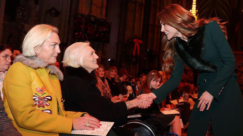 Annabel Bosher me të verdhë dhe Dame Mary Berry me të zeza janë të dyja ulur, ndërsa Princesha Catherine po i shtrëngon dorën Dame Mary Berry.