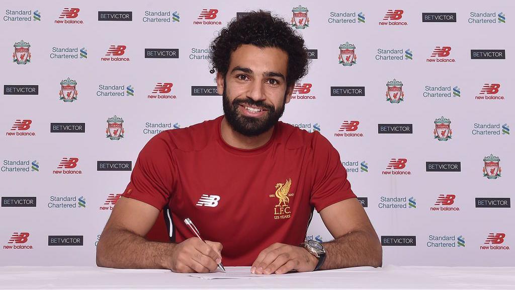 Mo Salah Signs a contract. He is holding a pen against the paper and is looking smiling into the camera. 