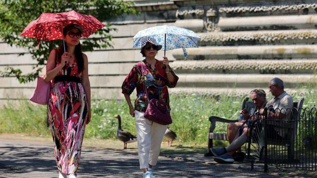 Two women walk in the sun, being shaded by umbrellas. They walk past a park bench, where a man and a women are sitting