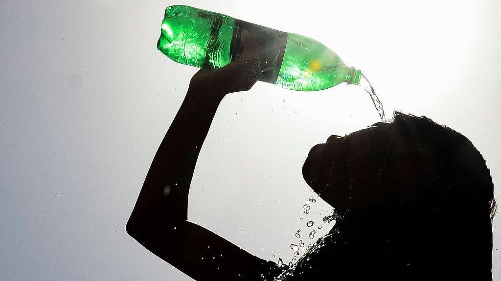 A young woman with long hair is silhouetted against the sun as she pours water on her face from a green plastic bottle held above her head. 