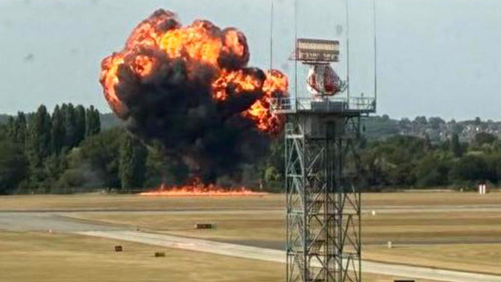A huge cloud of black smoke is rising into the air. There is fire on the ground and also filing the cloud of thick smoke. It is coming from the edge of an airfield with trees just behind it. The sky is clear blue, in stark contrast with the 'fireball'.