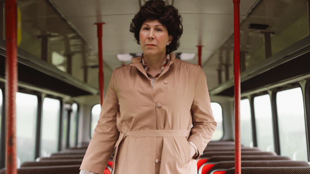 A woman stands in a long beige coat in the middle of the isle in a vintage bus. She had curled brown hair. 