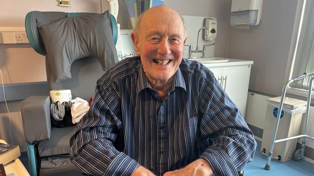 Robert Eaves wearing a striped blue and white shirt. He is smiling towards the camera. There is a grey chair in the background and a white sink unit on the right.