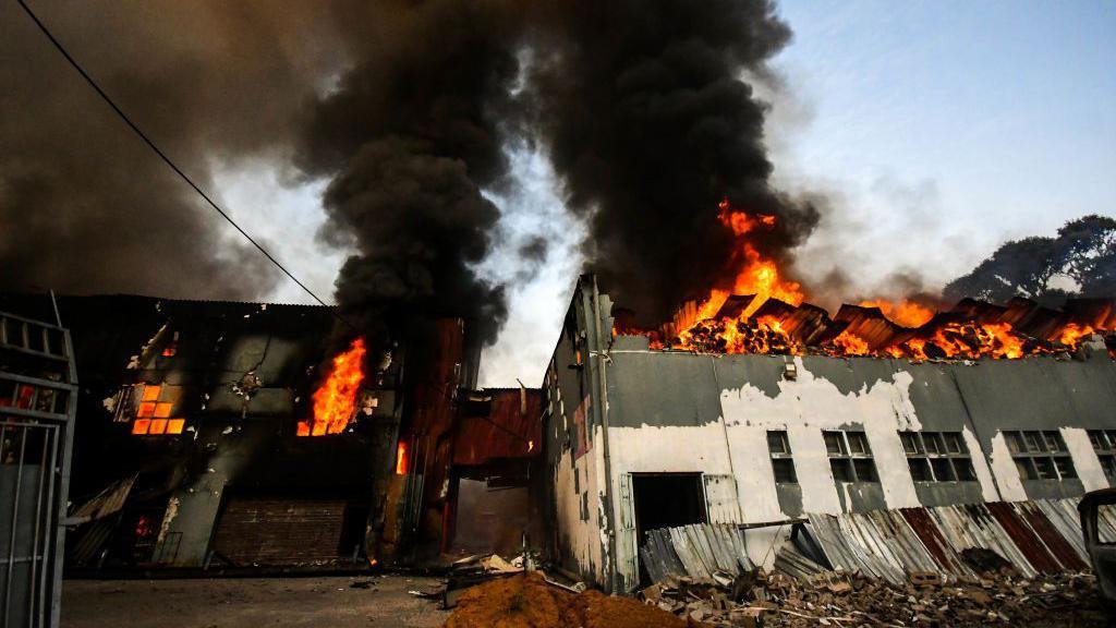 A factory in flames. Bright orange fire and thick black smoke can be seen coming from the top floor of the building. 