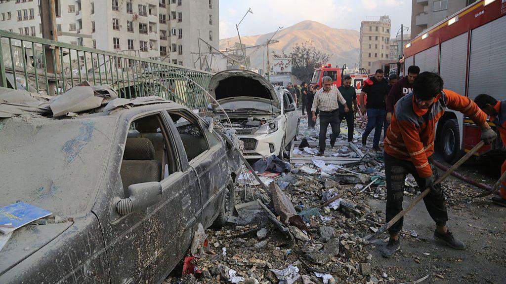 View of damaged building, person cleaning up rubble next to a car
