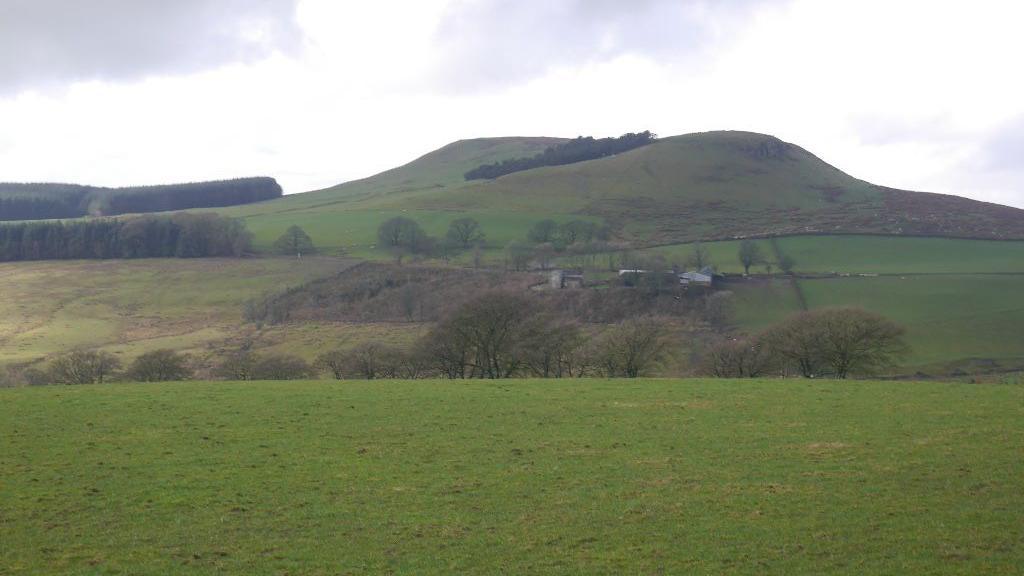 The site of a British hillfort near Lockerbie - a large mound of earth, completely covered with grass set in rural countryside with trees, fields and what looks like a farm building
