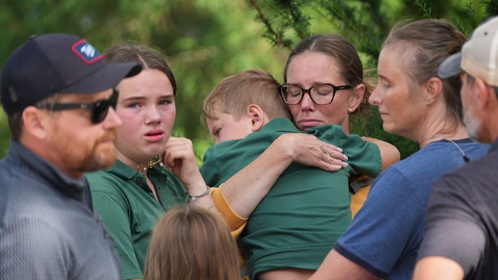 A group of parents and children stand together, looking emotional. One woman in glasses with her eyes closed hugs a young boy in a green shirt in her arms. Next to her, a teenage girl in a similar green shirt cries and clutches her necklace.