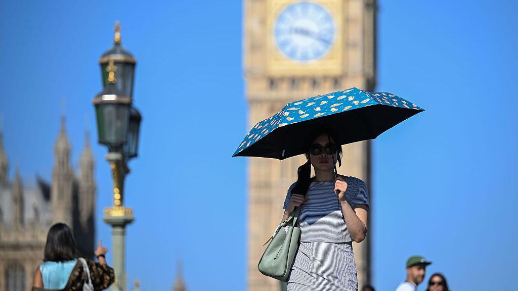 A woman walks in the shade of an umbrella in front of Big Ben in London, with clear blue skies overhead