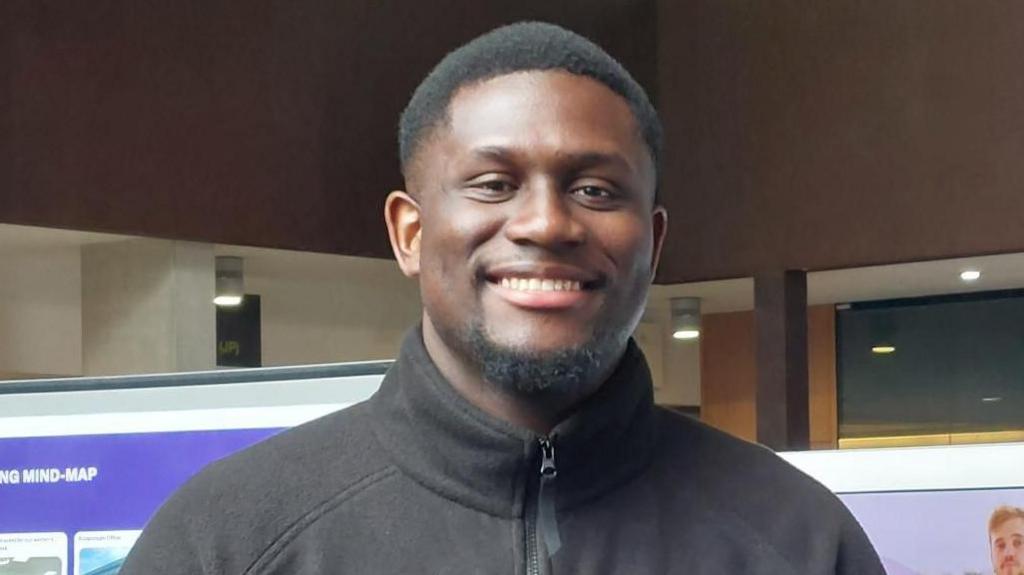 Kofi Owusu-Aidoo smiling for the camera at the Navigating Hybrid Work exhibition at Oxford Brookes University. Panels of the exhibition could be seen behind him.