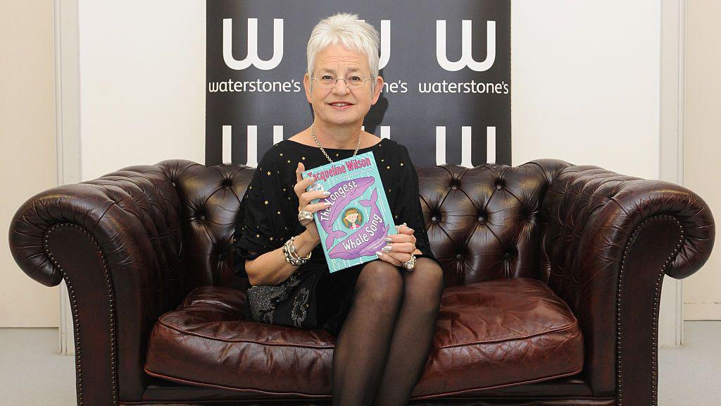 Jacqueline wilson sitting on a brown leather sofa posing with her book the longest whale song