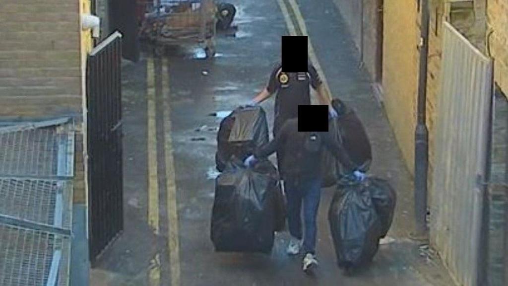 CCTV of two people carrying black bin bags down a back alley. Their faces have been obscured.