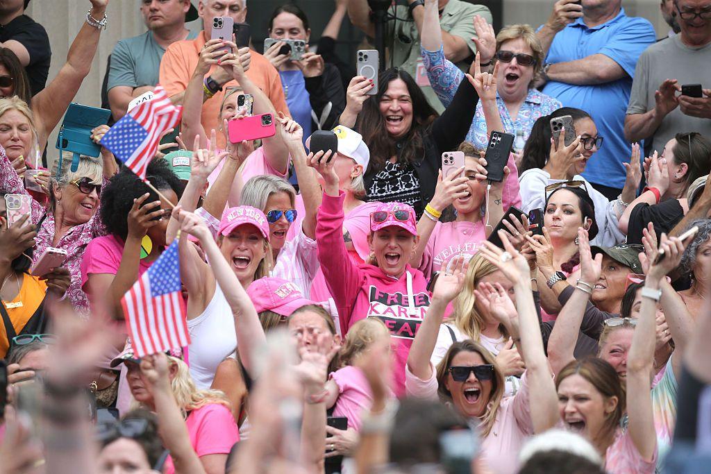 Supporters cheer outside court as Karen Read is acquitted of murder and manslaughter charges. Some are wearing pink shirts and hats and others are holding the US flag.