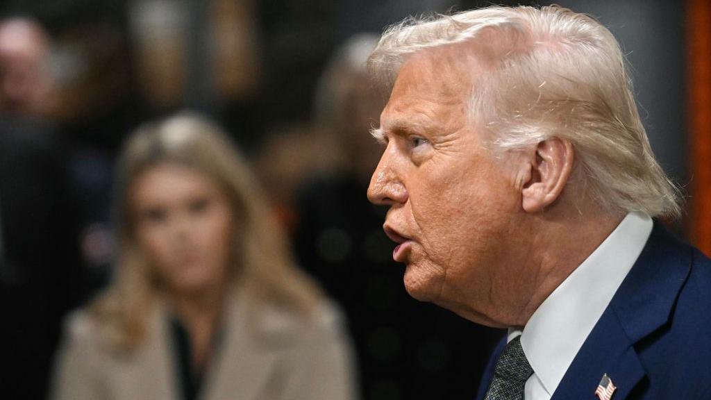 Donald Trump, in profile talks to members of the media as he visits the John F. Kennedy Center for the Performing Arts in Washington, DC, on Monday. There is a blurred figure of a young woman in the background.