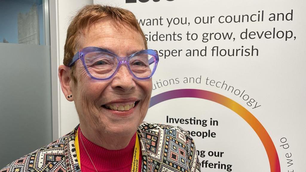 Conservative Councillor Carole Gandy, the cabinet member for adult social care at Herefordshire Council, is smiling at the camera - she is wearing a patterned jacket and a red jumper.