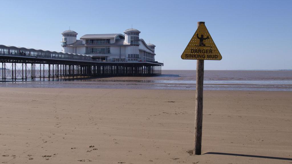 A triangular yellow warning sign on top of a signpost on Weston-super-Mare's beach, next to the pier. The signs says 'danger, sinking mud' with a silhouette image of a man up to his knees in the sand.