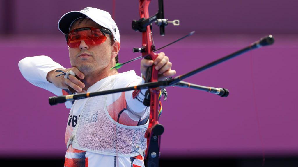 Tom Hall wearing a white cap and red sunglasses. He is holding a red recurve bow