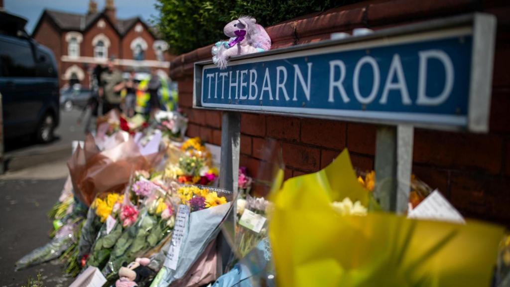 Bouquets of flowers on pavement in front of sign reading 'Tithebarn Road'
