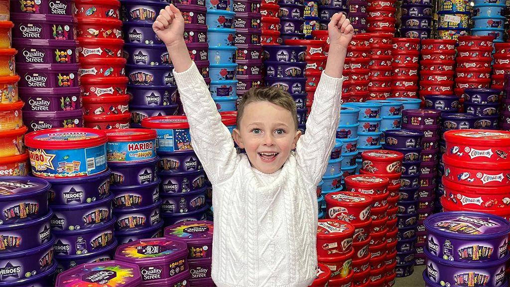 Teddy wears a white cable-knit jumper. He has short light brown hair. His arms are raised in triumph. He is in a warehouse full of thousands of sweet tubs, including Heroes, Quality Street, and Celebrations.