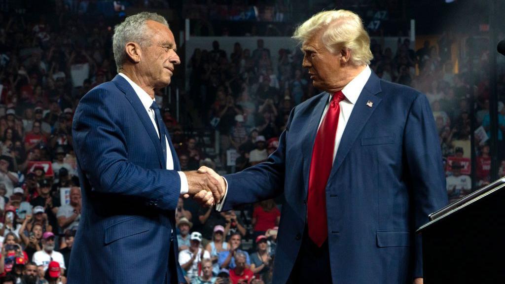 Robert F Kennedy in a blue suit with blue tie shaking hands with Donald Trump in a blue suit with red tie