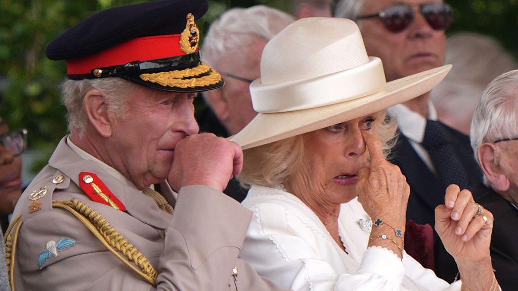 King Charles III and Queen Camilla are seated, with the Queen dabbing her eyes, apparently tearful, and the King visibly emotional. The King is dressed in military uniform, while the Queen wears white clothing and a hat