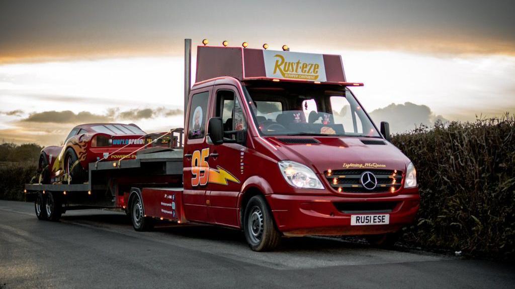 A large transite van with a flatbed, carrying a car on the back. It is on a road next to a hedgerow. The van is burgundy and the car on the back is bright red.