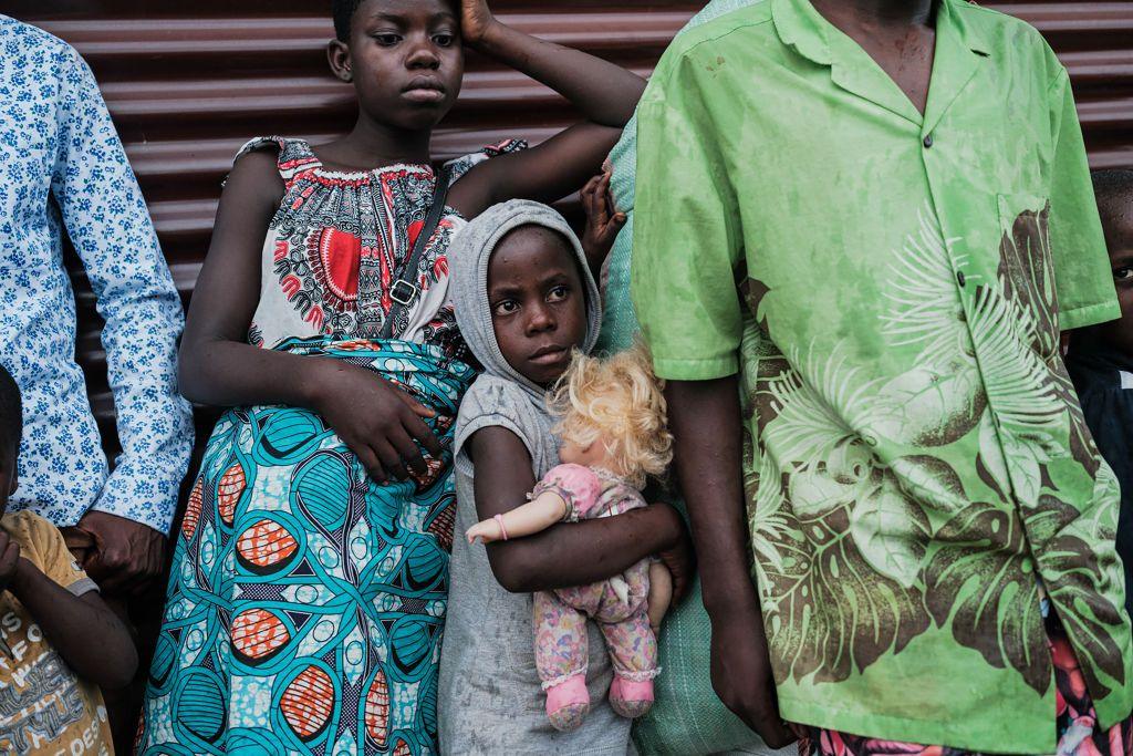 A little girl is pictured holding a doll as she lines up with her family at a refugee camp. The girl looks serious and worried. An older girl beside her rests against a corrugated metal fence. 